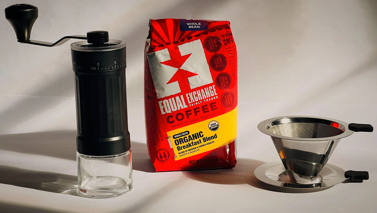 A manual black and clear coffee grinder, a red and yellow bag of Equal Exchange Coffee, and a stainless steel pour-over dripper on a white background with dramatic shadows.