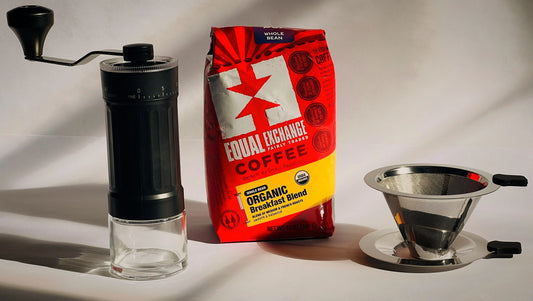 A manual black and clear coffee grinder, a red and yellow bag of Equal Exchange Coffee, and a stainless steel pour-over dripper on a white background with dramatic shadows.