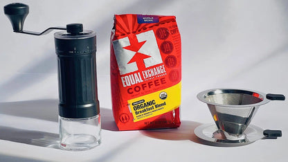 A coffee brewing set on a white surface with strong shadows, featuring a manual black-and-clear grinder, a red and yellow bag of Equal Exchange coffee, and a stainless steel pour-over filter with a metal tray.