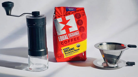 A coffee brewing set on a white surface with strong shadows, featuring a manual black-and-clear grinder, a red and yellow bag of Equal Exchange coffee, and a stainless steel pour-over filter with a metal tray.